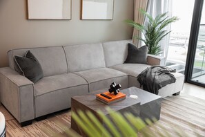 A modern living room with a grey sofa, a coffee table with a book and a decorative object, and a large window with a view of the city. - StayShort - Chic 1 Bedroom Unit in Dubai Marina that Sleeps 4! (Dubai)
