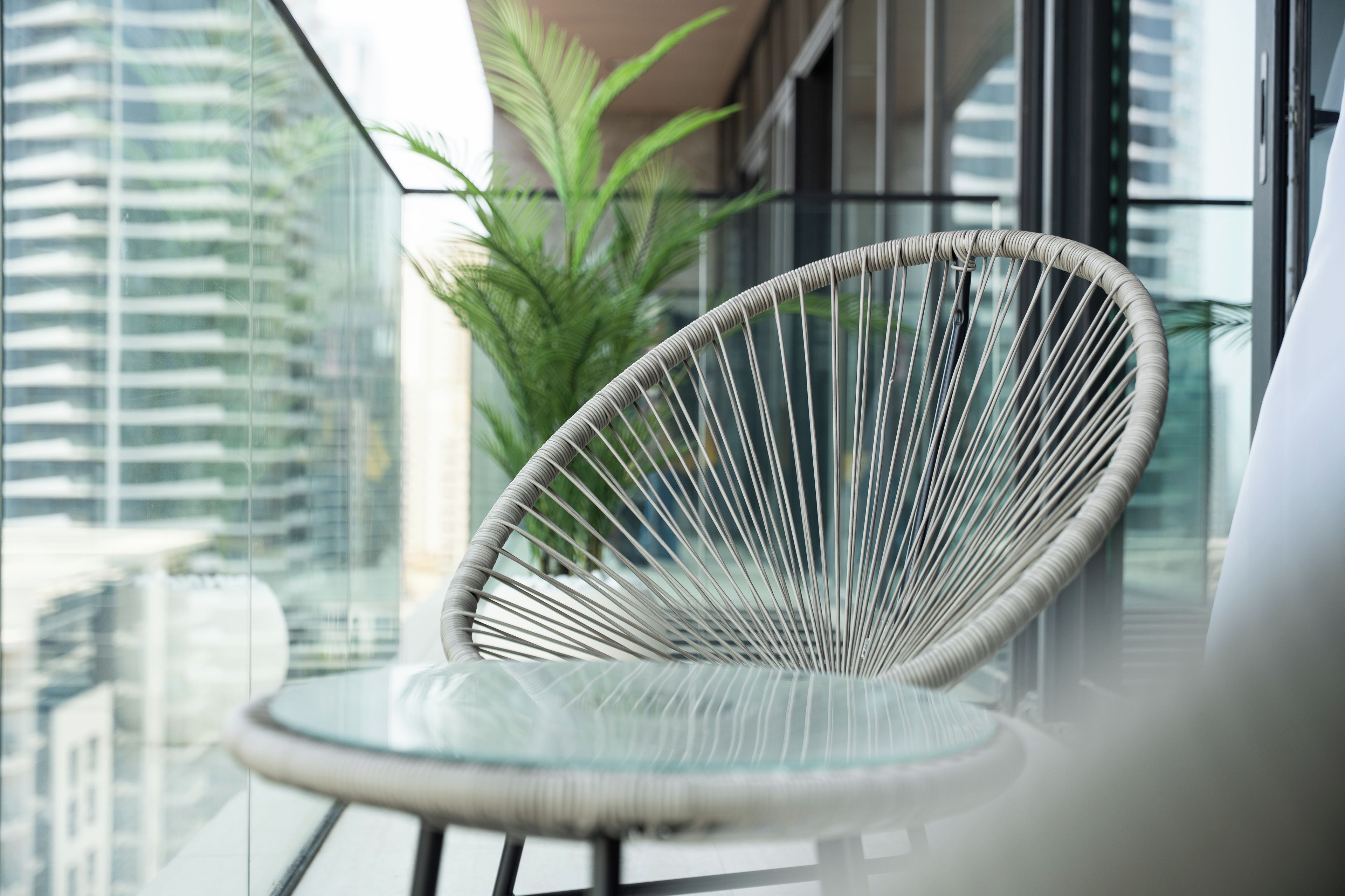 A wicker chair with a glass table on a balcony with a city view.