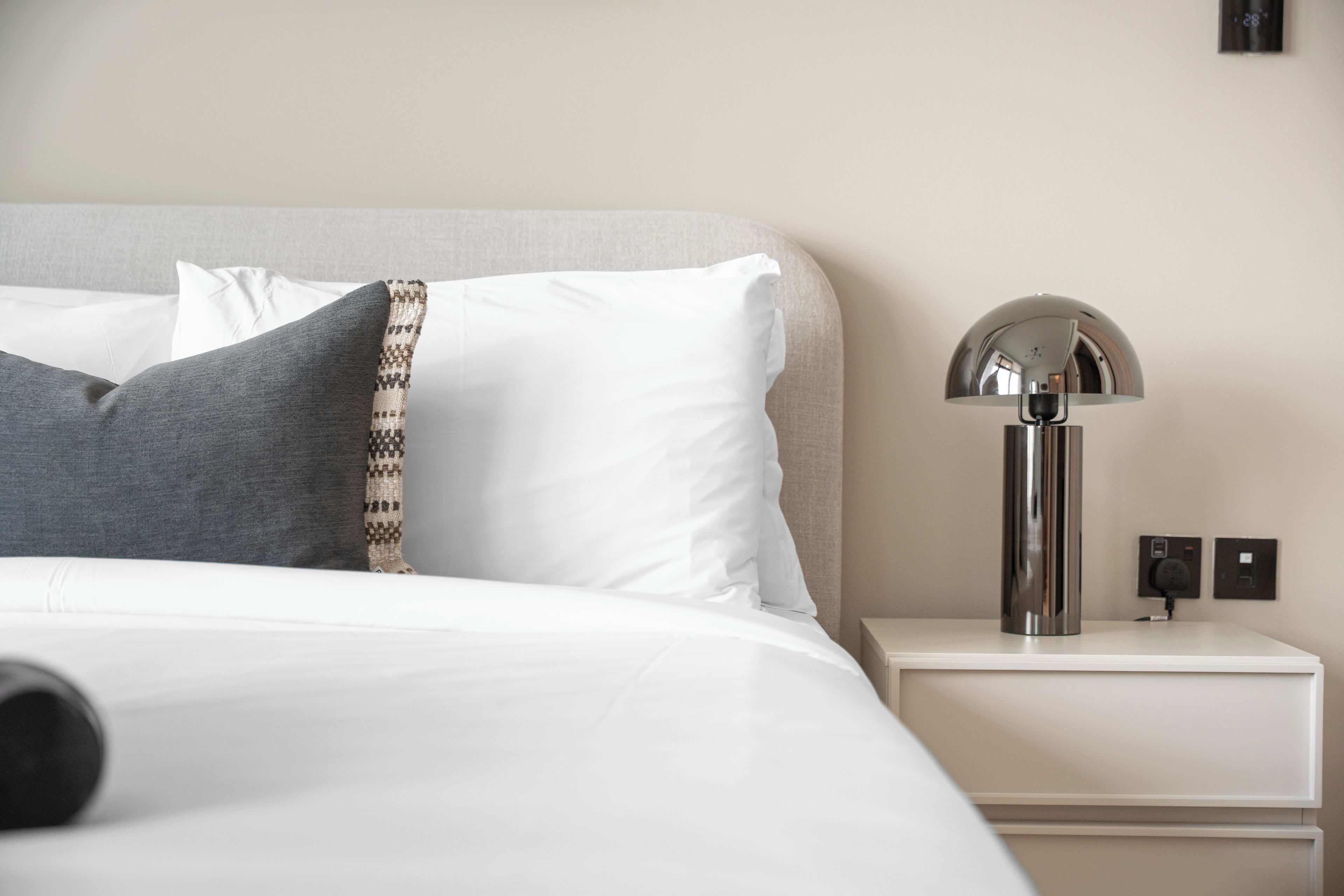 Un lit bien fait, avec des oreillers blancs et gris, un plaid à motifs et une lampe moderne posée sur une table de chevet.