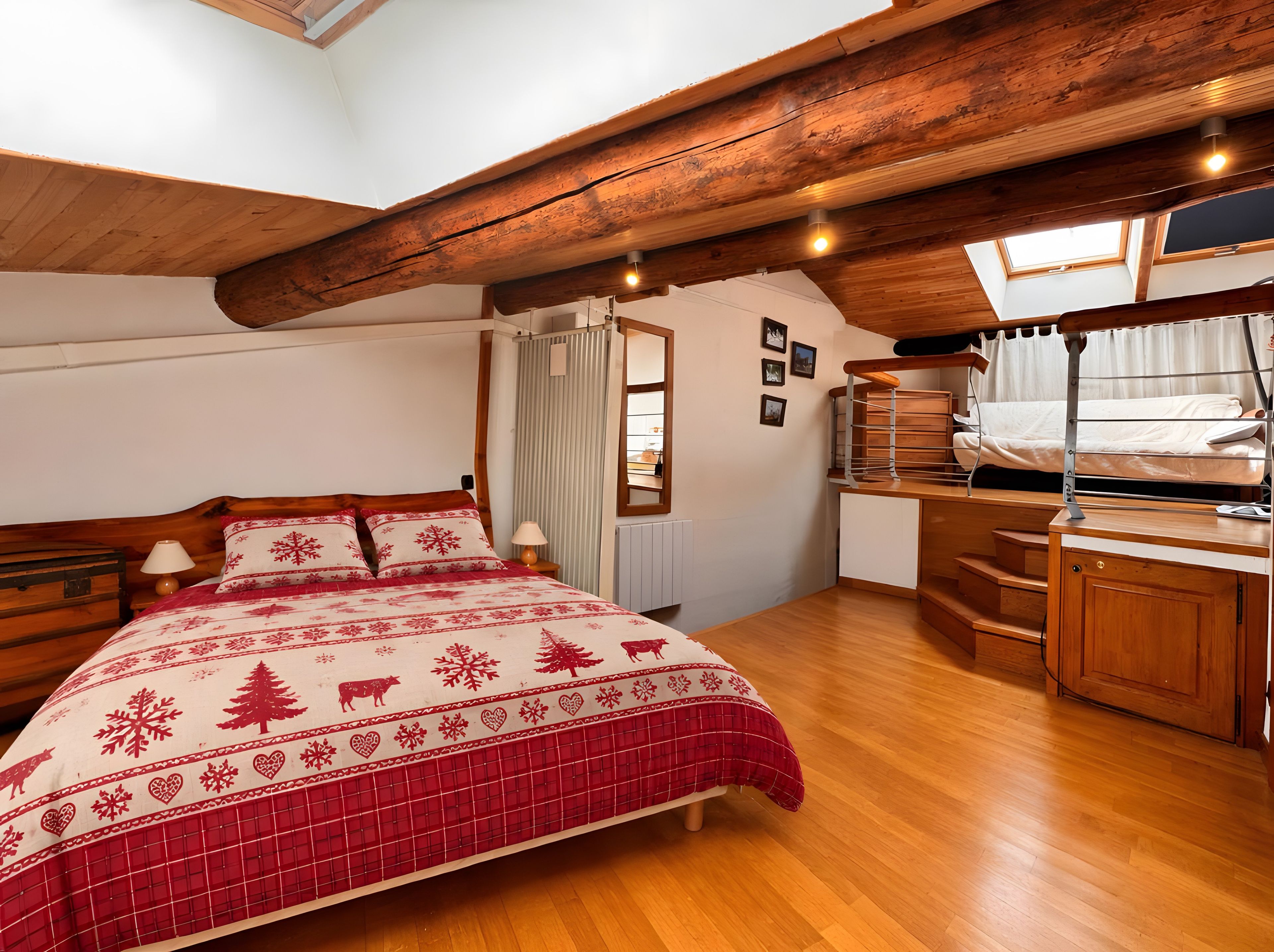 A cozy bedroom with a wooden ceiling, a bed with a red and white patterned bedspread, a desk, and a window with curtains.