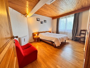 Un dormitorio con techo de madera, una cama, una silla roja y una ventana con cortinas.