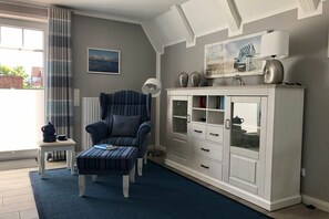 Ein Zimmer mit einem blauen Sessel, einem weißen Schrank mit Glastüren, einem kleinen Tisch mit einer Lampe und einem Fenster mit Vorhängen.