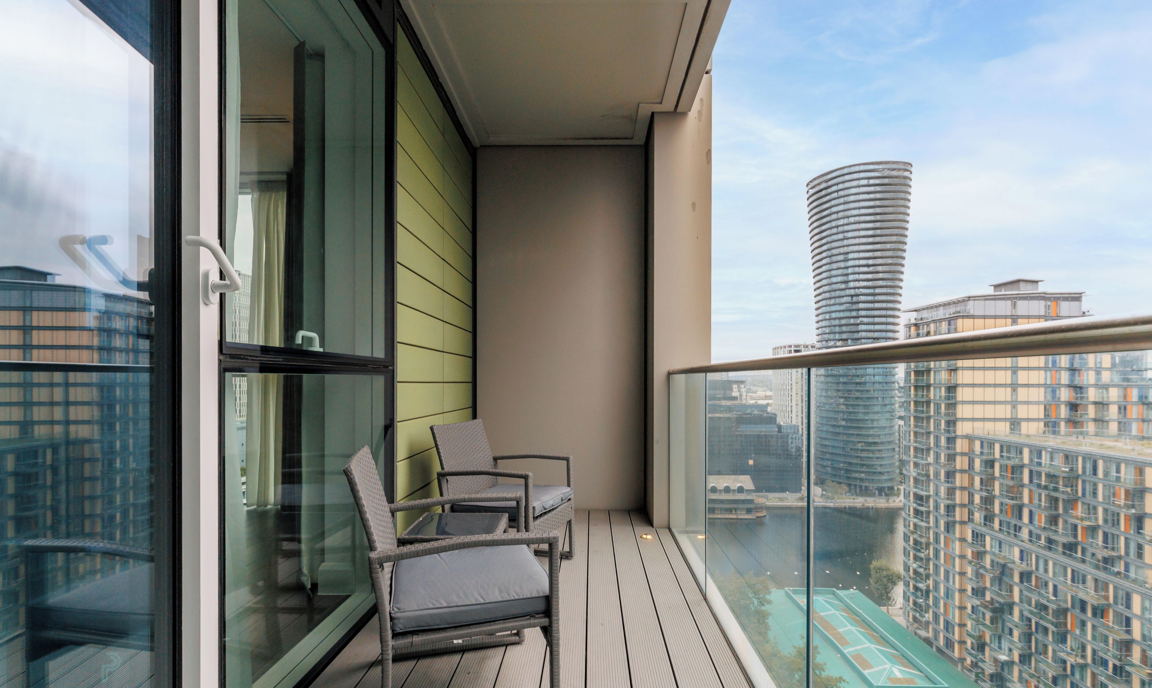 A balcony with a glass railing, a chair, and a view of a cityscape.