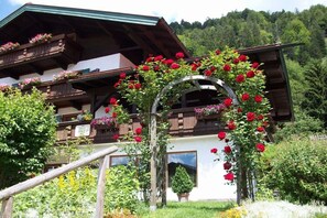 Ein traditionelles Chalet mit Holzbalkonen, einem hölzernen Bogen und einem Garten mit roten Blüten.