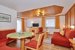 Ein möbliertes Wohnzimmer mit Holzbalkendecke, einem Esstisch mit Stühlen, einer Couch und einem Fernseher.