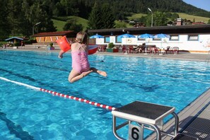Ein Schwimmbecken mit Sprungbrett, einem Rettungsschwimmerstuhl und einem Gebäude mit Außenbereich im Hintergrund.