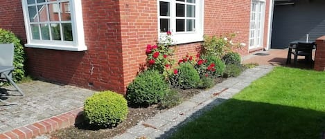 A brick house with two windows, a garage, and a small garden with red flowers and shrubs.