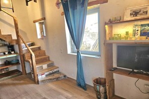 A staircase with wooden steps and a blue curtain, a television on a wooden shelf, and a window with a view of the outside. - Agio Farago Gallery Studio (Pitsidia)