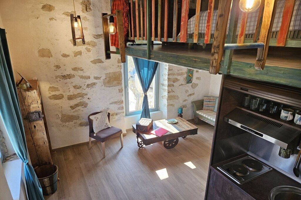 A loft-style room with a loft bed, a small table with a chair, a window with curtains, and a kitchenette area.