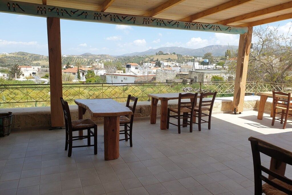 An outdoor patio with wooden tables and chairs, a tiled floor, and a view of a village and hills in the distance.