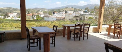 An outdoor patio with wooden tables and chairs, a tiled floor, and a view of a village and hills in the distance.