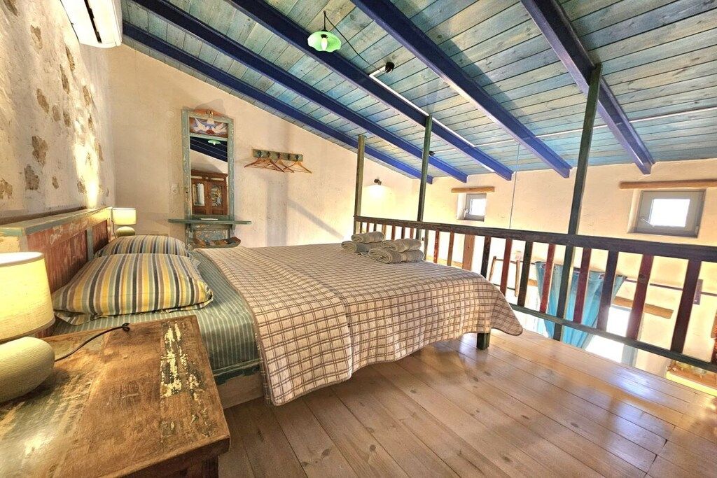 A bedroom with a wooden floor, a bed with a plaid bedspread, a nightstand with a lamp, and a window with a view.