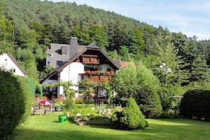 A two-story house with a balcony, surrounded by a well-maintained garden and a forested hillside. - Haus Winterberg || Erfweiler (Erfweiler)