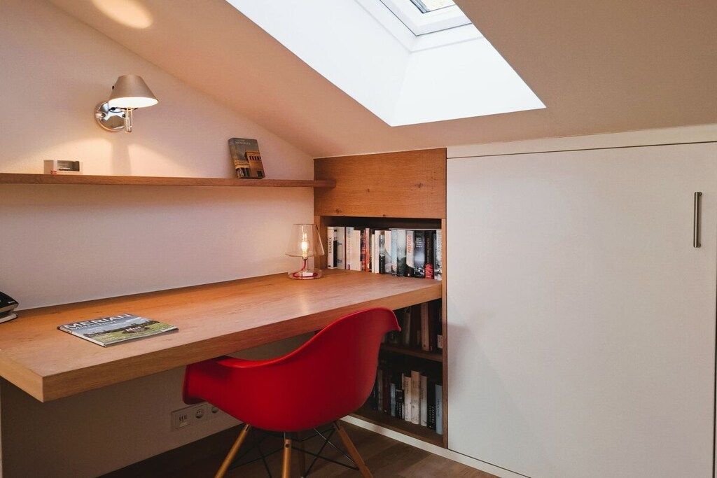 Una habitación pequeña con tragaluz, un escritorio de madera, una silla roja, una estantería y un armario blanco.