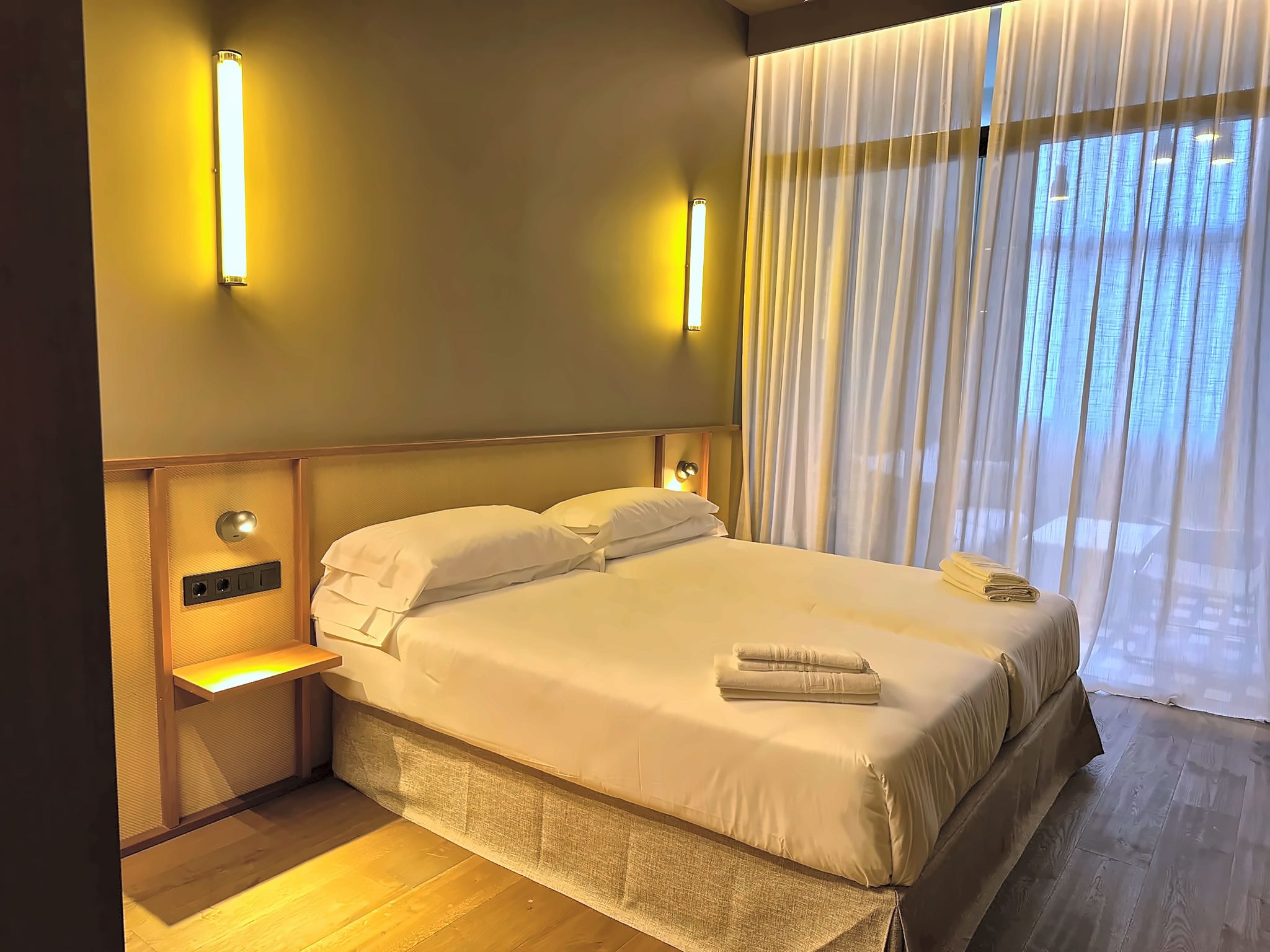 A hotel room with a bed, bedside lamps, a nightstand, and a window with curtains.