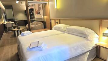 Habitación de hotel con una cama bien hecha, una mesita de noche con lámpara, un espejo y una zona de estar con una silla y una mesa.