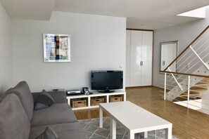 A modern living room with a grey sofa, a flat-screen TV on a white stand, a staircase with a wooden railing, and a framed picture on the wall. - Holiday home with 2 bedrooms (Seebad Heringsdorf)