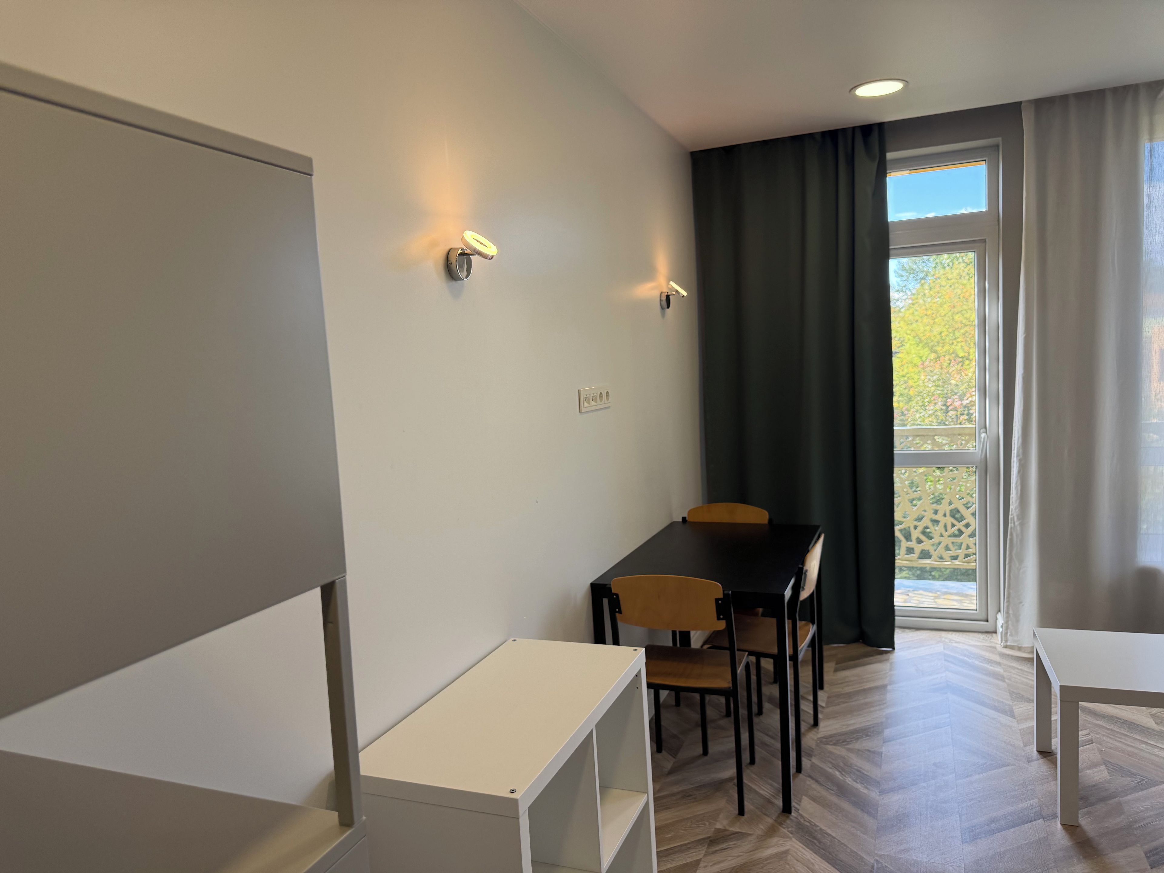 A modern, well-lit room with a dining table and chairs, a small white desk, and a window with curtains.