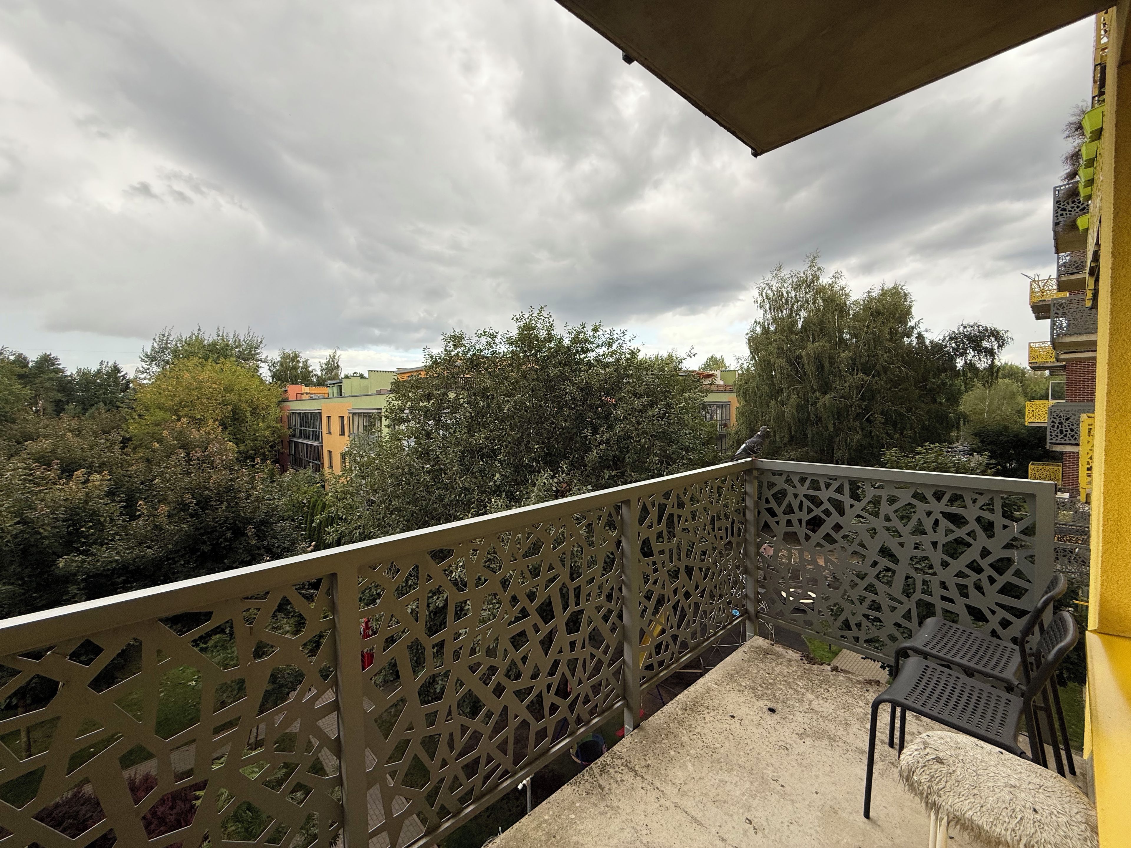 A balcony with a lattice railing, a concrete floor, and a view of surrounding buildings and trees.
