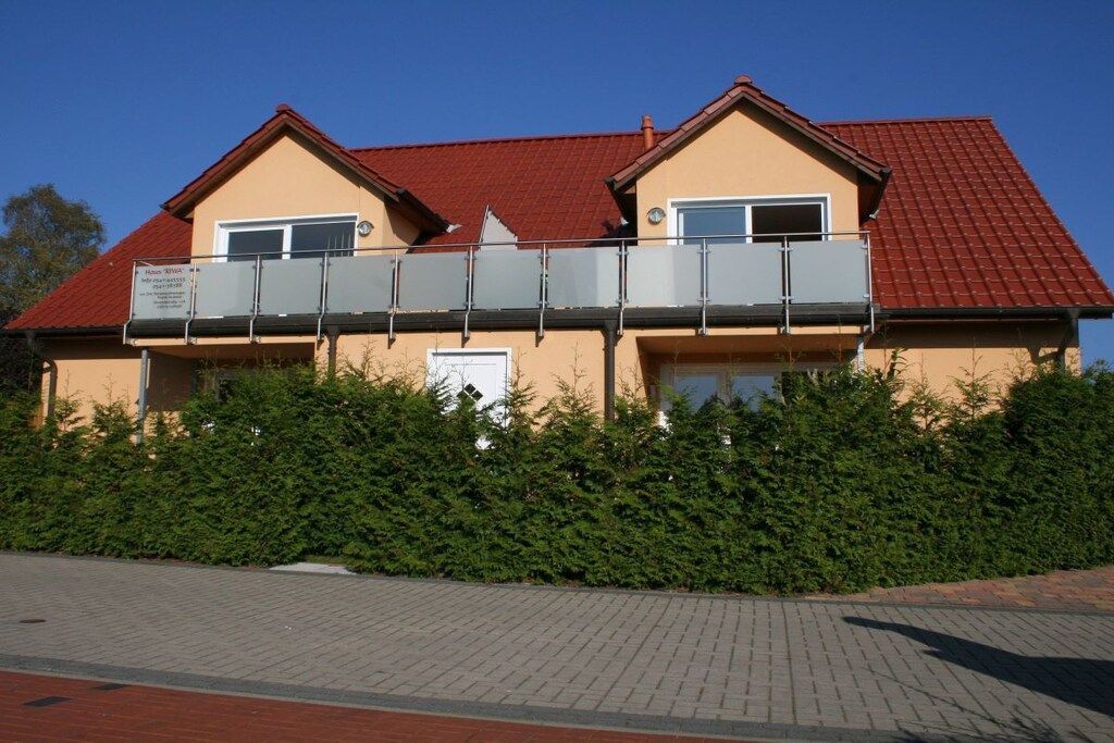 A two-story building with a red roof, a balcony, and a hedge in front.