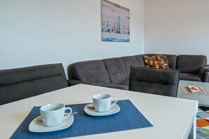 A room with a white table, two white cups on saucers, a blue placemat, a grey sofa, and a colorful rug. - Holiday apartment with 1 bedroom (Bensersiel)