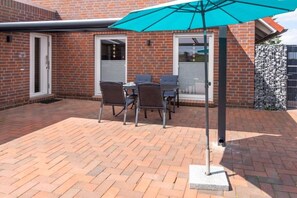 Een terras met een geplaveid vloertje, een tafel met stoelen en een blauwe parasol.