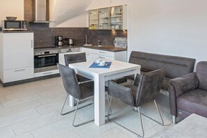 A compact kitchen with modern appliances, a dining area with a white table and grey chairs, and a grey sofa. - Holiday apartment with 1 bedroom (Bensersiel)