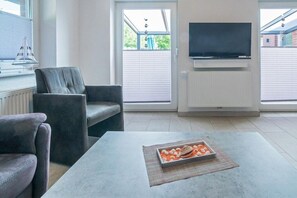 A modern living room with a sofa, a coffee table with a decorative item, a television mounted on the wall, and a window with blinds. - Holiday apartment with 1 bedroom (Bensersiel)