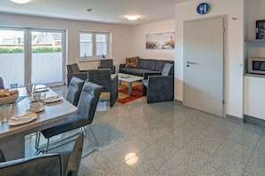 A modern living room with a dining area, grey sofas, a coffee table, and a door with a sign. - Holiday home with 1 bedroom (Bensersiel)