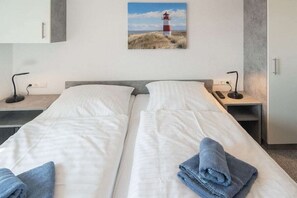 A double bed with white linens, two bedside tables with lamps, a painting of a lighthouse, and a small shelf. - Holiday home with 1 bedroom (Bensersiel)