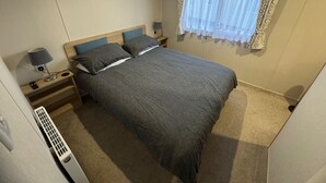 A bedroom with a bed, bedside lamps, a heater, and a window with curtains. - 2-bed - Sleeps 4 - Free Parking (Sandown)