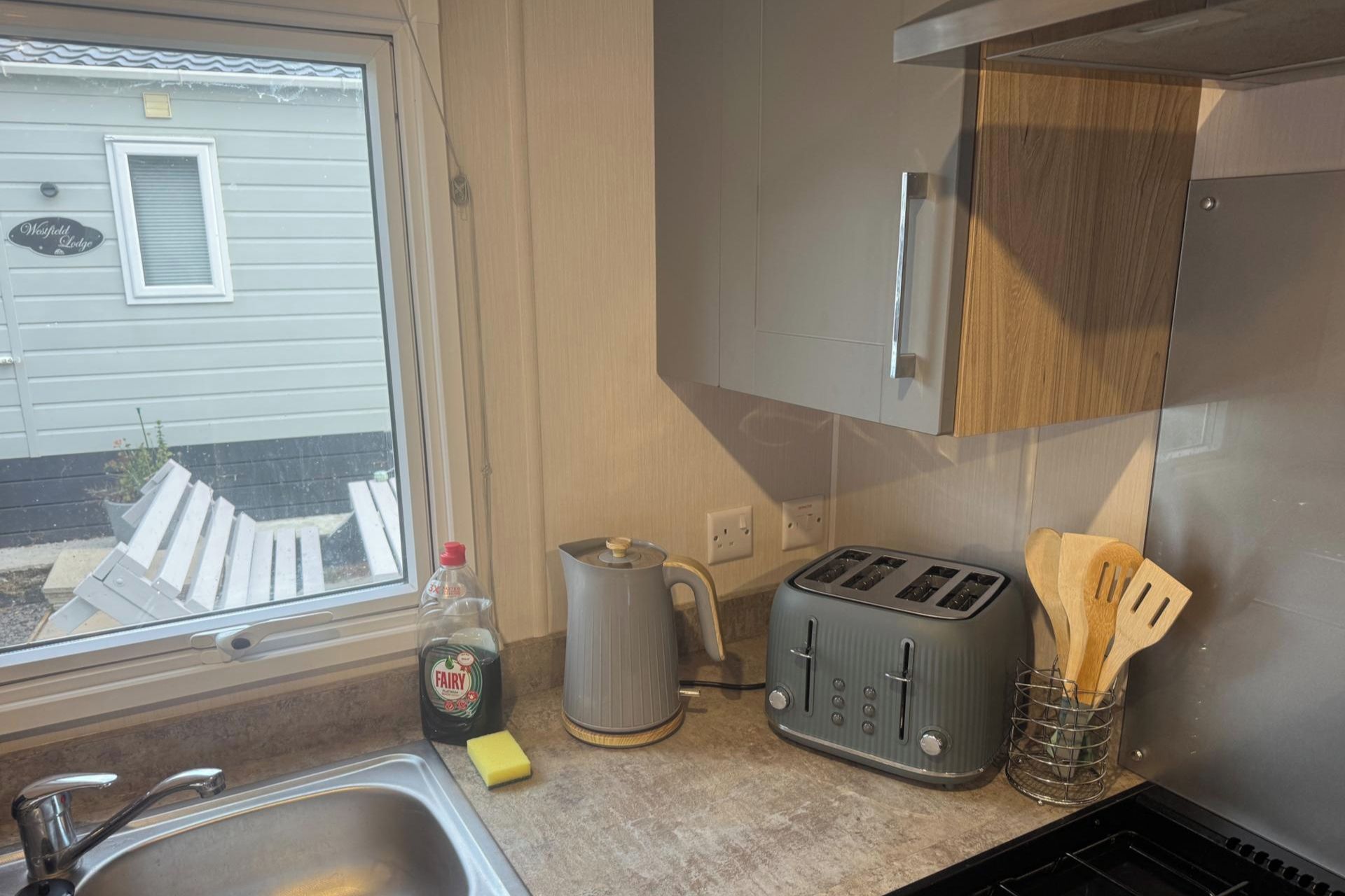 A kitchen with a sink, a toaster, a kettle, and a window with a view of a house.