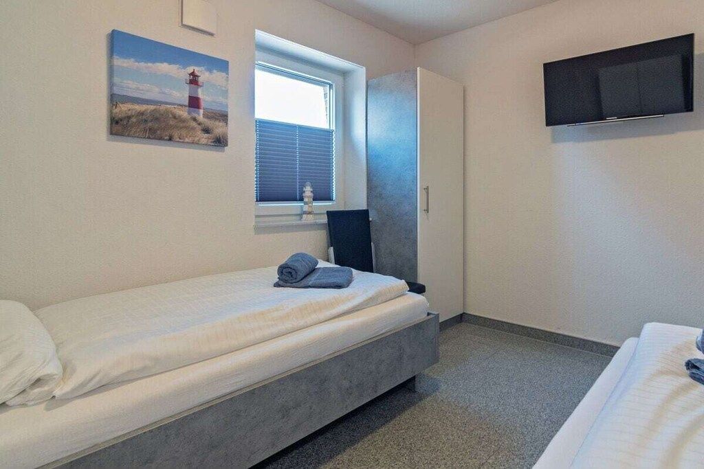 A single-bed room with a window, a flat-screen TV mounted on the wall, and a painting of a lighthouse.