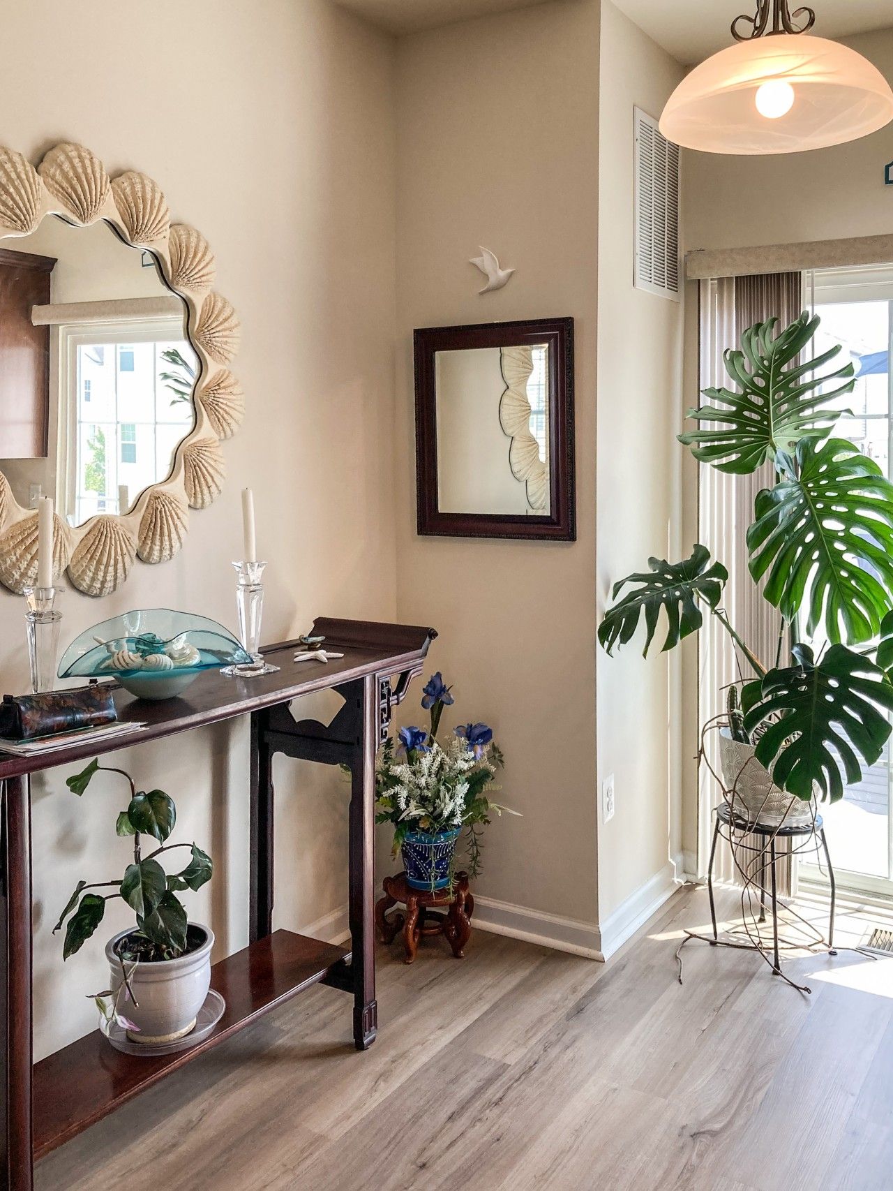 A room with a wooden dining table, a mirror with a shell frame, a potted plant, and a hanging light fixture.