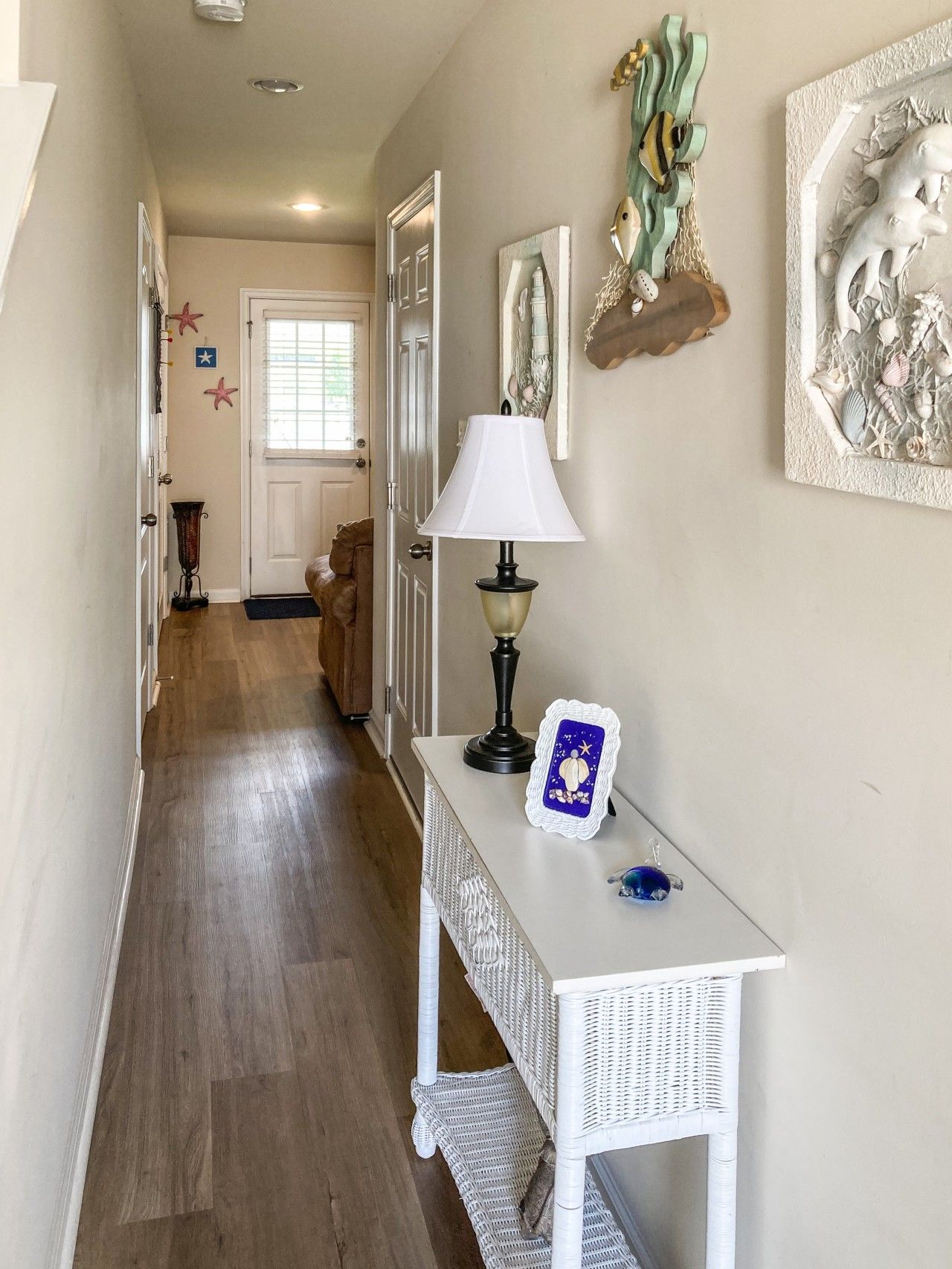 Um corredor com um console de vime branco, uma luminária e quadros nas paredes.