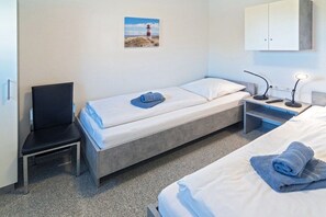 A single bed with a pillow and a rolled towel, a chair, a bedside table with a lamp, and a wall-mounted cabinet. - Holiday home with 1 bedroom (Bensersiel)