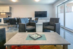 A modern living area with a flat-screen TV, seating, and a kitchen in the background. - Holiday home with 1 bedroom (Bensersiel)