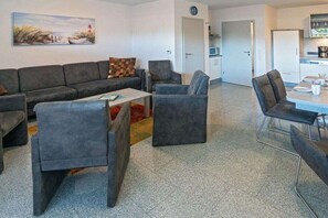 A modern living room with a sofa, armchairs, and a coffee table. There is a kitchen area with cabinets and a refrigerator. - Holiday home with 1 bedroom (Bensersiel)