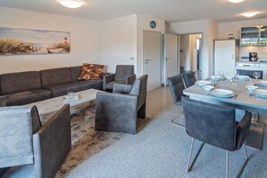 Un salon moderne comprenant un canapé, des fauteuils, une table basse et un coin repas avec une table dressée pour quatre personnes.