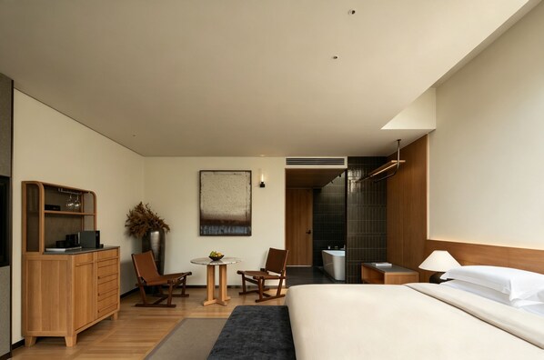 A modern hotel room with a bed, a desk, a chair, a small table, and a shower area.