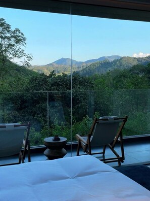 A room with a bed, two chairs, and a small table, overlooking a mountainous landscape.