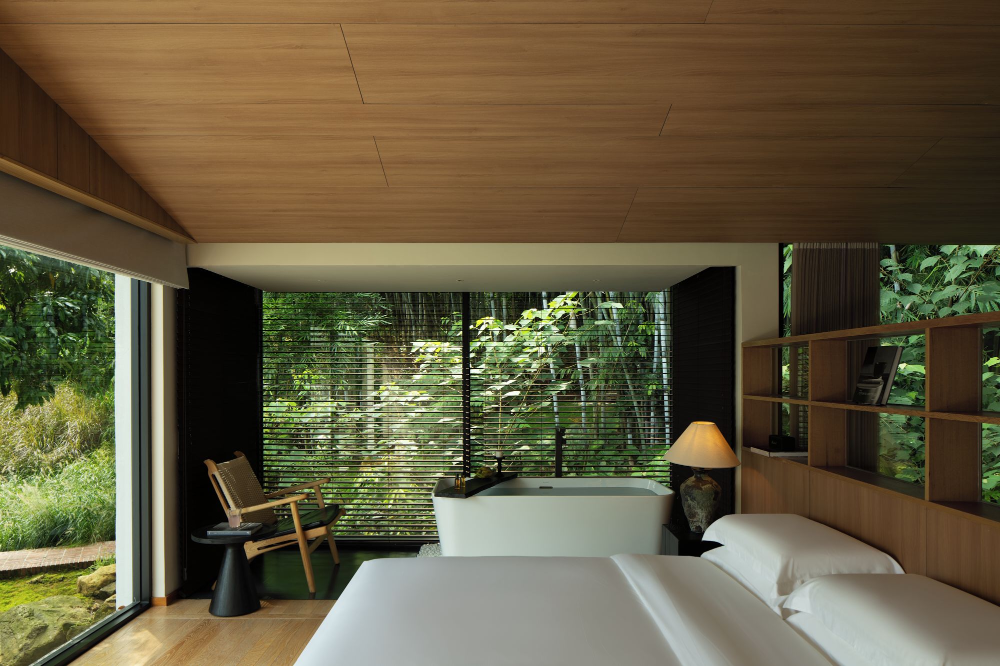 Une chambre moderne avec un grand lit, une table de chevet, une chaise et une vue sur la verdure par la fenêtre.