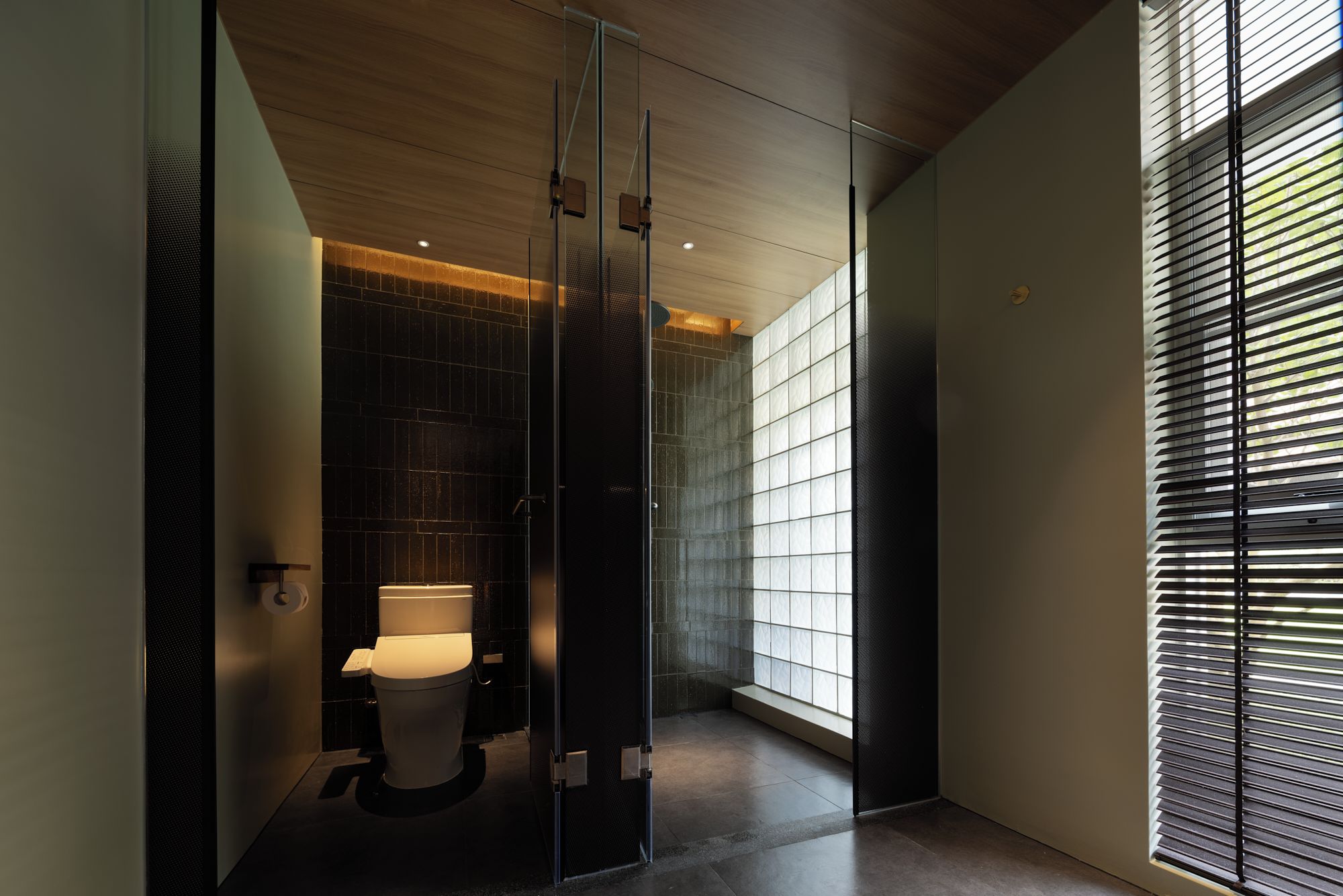 A modern bathroom with a glass shower enclosure, a toilet, and a window with blinds.