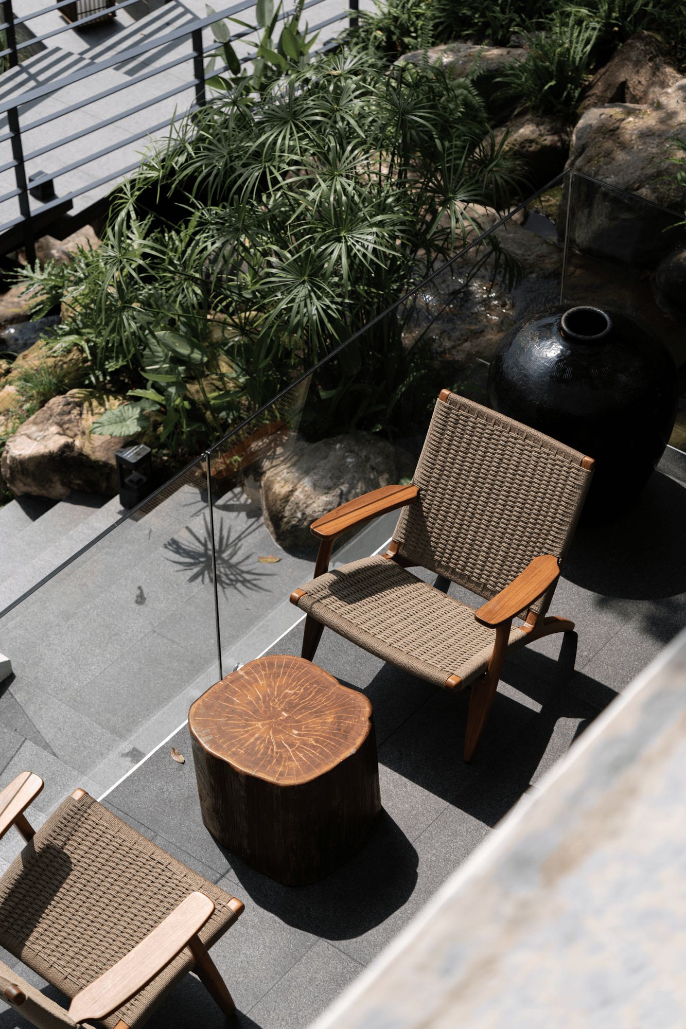 Una silla de mimbre y una mesa redonda de madera en un espacio de estar al aire libre, con rocas y plantas de fondo.