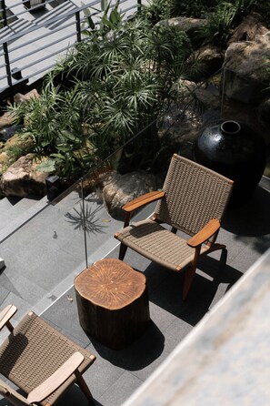 A wicker chair and a round wooden table in an outdoor seating area with a rock and plant backdrop.