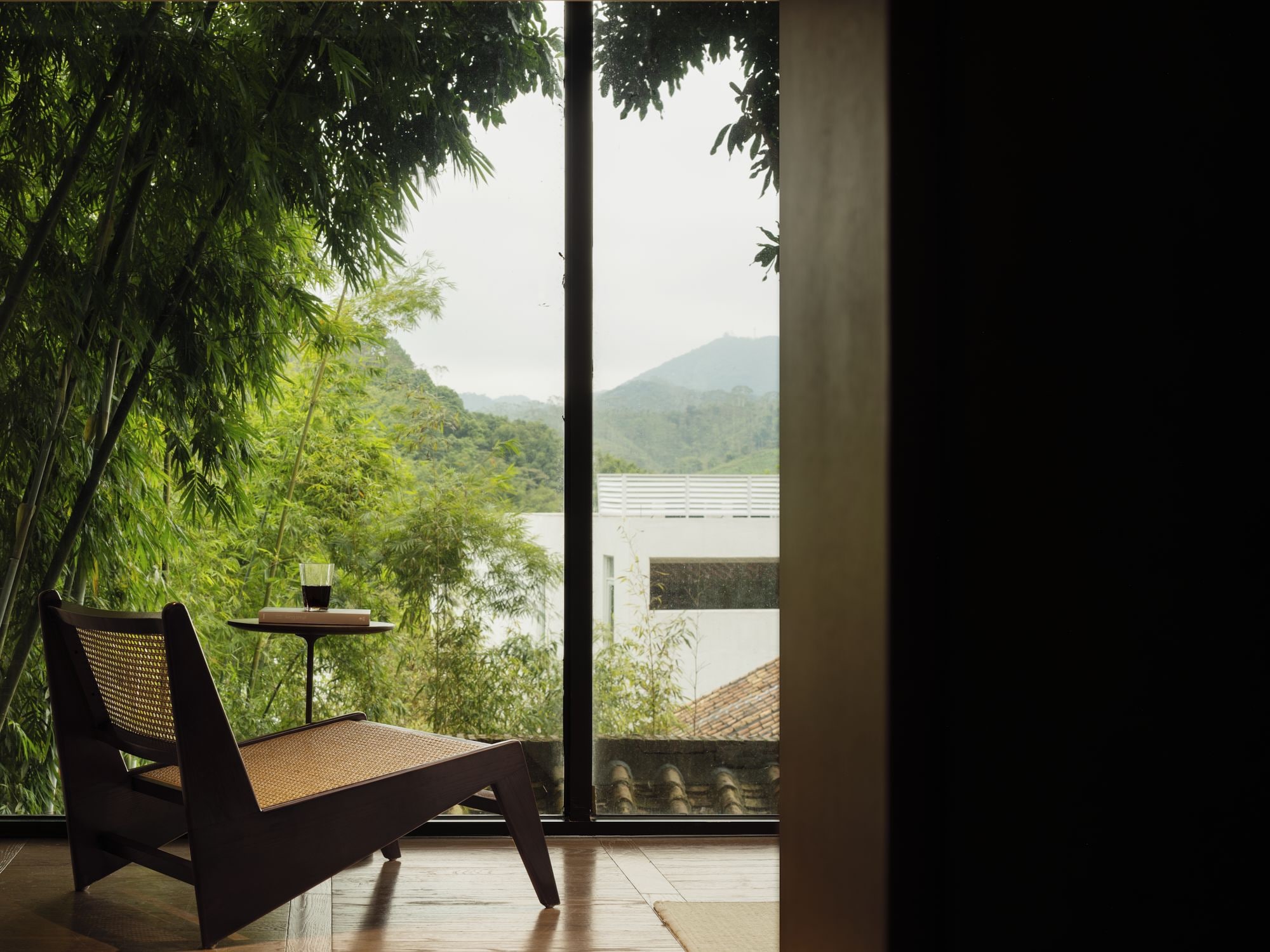 Una habitación con un gran ventanal que da a un paisaje exuberante, un sillón de mimbre y una mesita con un vaso de bebida.