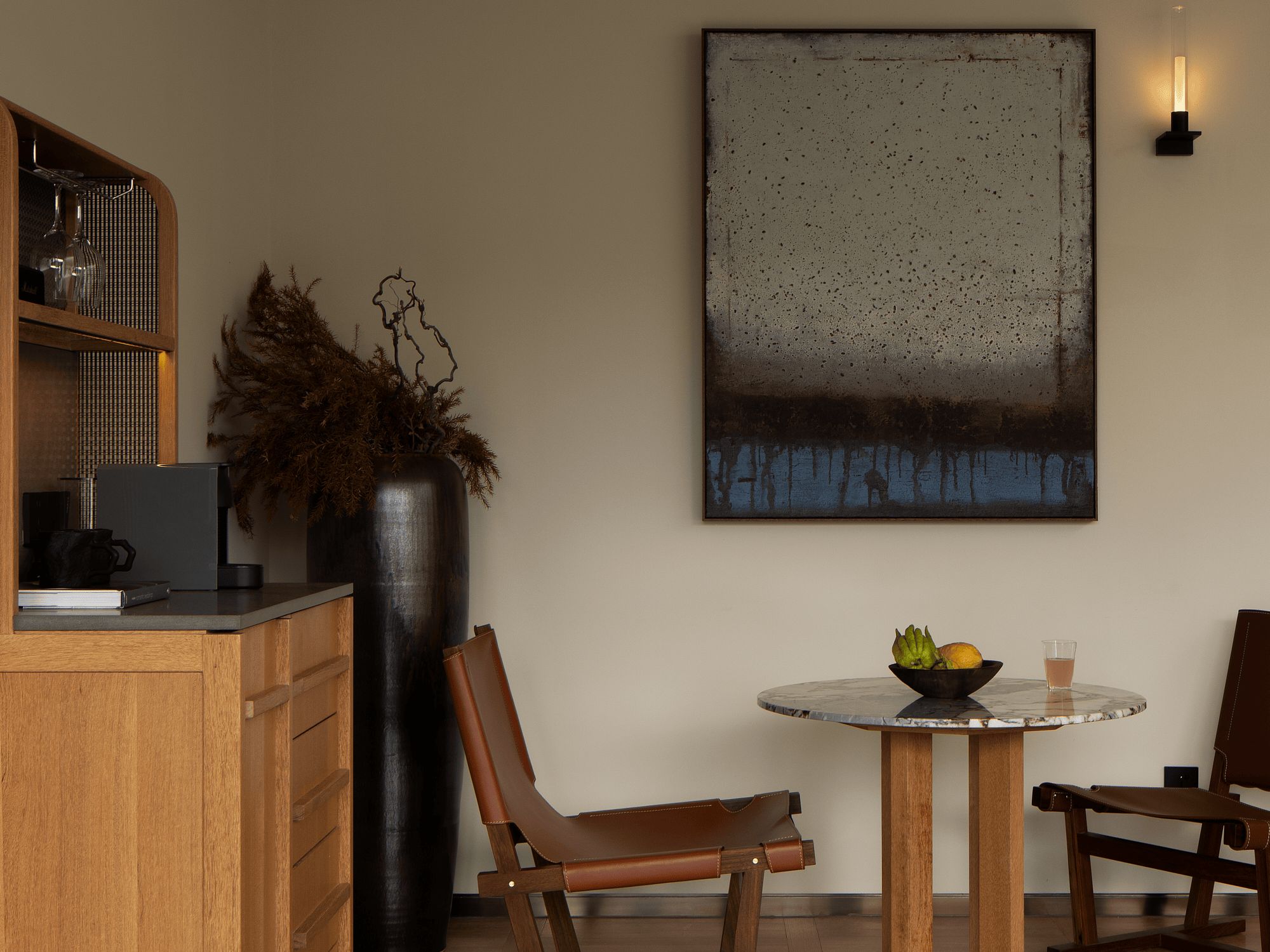 Una cocina moderna con comedor, que presenta un cuadro abstracto grande, un florero negro con ramas secas y una mesa redonda con un cuenco de fruta.