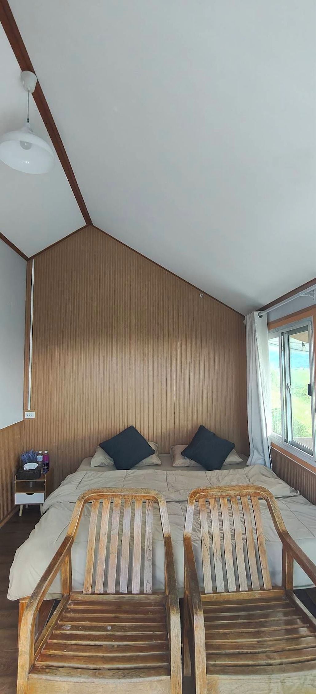 A room with a sloped ceiling, a bed with two pillows, a nightstand with a lamp, and two wooden chairs.