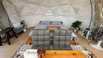 Interior acogedor de una tienda en forma de cúpula con cama, zona de estar y una mesa con un ventilador y una botella de agua.