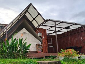 Una estructura triangular parecida a una tienda de campaña, con un banco de madera y una pequeña mesa, rodeada de follaje y plantas en macetas.